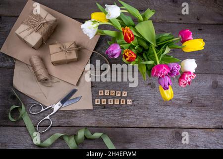 Frohe Ostereinschrift in Holzbuchstaben. Geschenkverpackung. Mix aus Frühlingsblumen. Hintergrund mit Blumen Tulpen close-up verschiedenen Farben. Geschenk. Draufsicht. Speicherplatz kopieren. Handgemacht Papper. Stockfoto