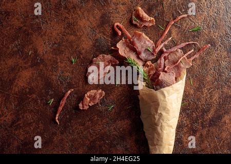 Scheiben von würzigem Trockenfleisch mit Rosmarin in einem Papierbeutel auf einem alten braunen Hintergrund. Draufsicht. Stockfoto