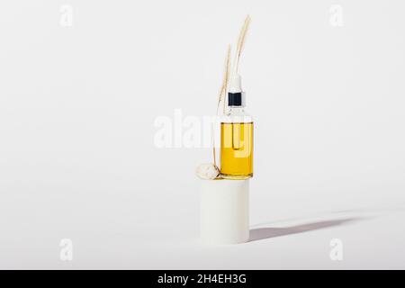 Naturkosmetiköl-Glasflasche mit Pipette steht auf dem Podium neben Stein und trockenem Gras auf weißem Hintergrund mit Kopierraum Stockfoto