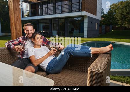 Glückliches romantisches Paar, das sich ausruhte und Wein trank, während es in der Nähe des Schwimmbades auf dem Land saß Stockfoto