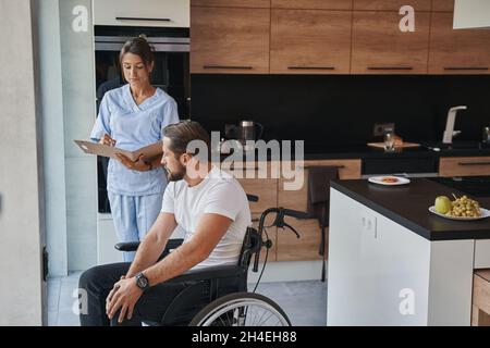 Schöner Arzt in Uniform und männlich, die mit einem Rollstuhl Gespräch in der Küche zu Hause Stockfoto