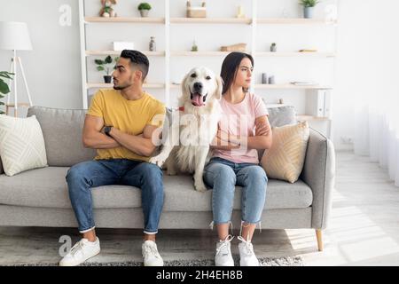 Unglücklich beleidigte das junge, vielfältige Paar, das in entgegengesetzte Richtungen schaute und auf dem Sofa saß, mit einem niedlichen Hund dazwischen Stockfoto