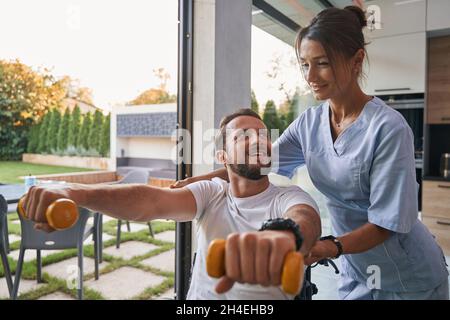 Entzückender kaukasischer Mann, der die Hantel in den Händen hält, während er auf die Physiotherapeutin im Zimmer schaut Stockfoto