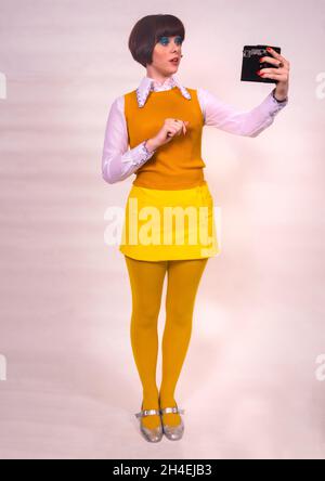 Die hübsche junge Frau im Stil der 1960er Jahre hält eine Kamera aus, als würde sie ein Selfie machen Stockfoto