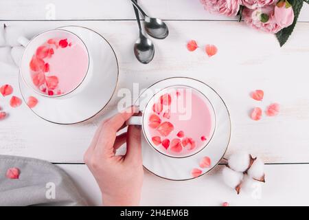 Rosa Matcha mit Rosenblättern auf weißem Hintergrund. Weibliche Hand hält eine Tasse mit trenderem veganem Tee. Entspannender Drink in der erholsamen Zeit. Stockfoto