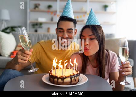 Ein junges, vielfältiges Paar mit Partyhüten feiert Geburtstag, bläst Kerzen auf den Festkuchen, trinkt Champagner zu Hause Stockfoto