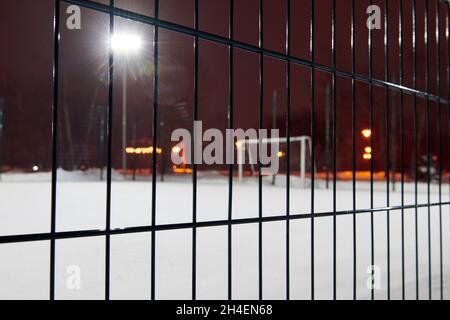 Fußballplatz mit einer dicken Schneeschicht bedeckt. Blick durch den Zaun. Konzentrieren Sie sich auf den Zaun. Verschwommenes Foto. Nachtspiel wird abgesagt Stockfoto