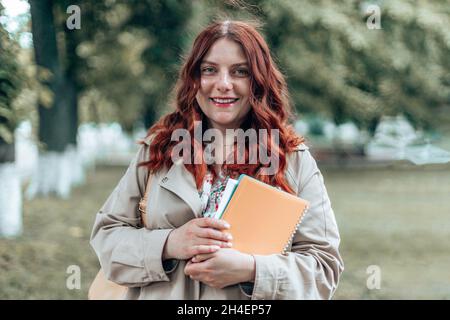 Junge kaukasische Studentin der Universität, die vor dem Campus eine Brille mit Büchern trägt. Feuriges lockiges Haar Stockfoto