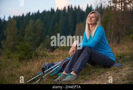 Schöne Frau mit touristischen Rucksack sitzt auf Gras in der Nähe des Waldes, um die Landschaft während der Sommerwanderung Abenteuer in den Bergen zu bewundern. Konzept von Reisen, Wandern und aktiver Freizeit. Stockfoto