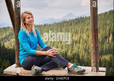 Seitenvide einer charmanten jungen Frau, die wegschaut und lächelt, während sie auf einer hölzernen Schaukel in den Bergen sitzt. Weibliche Reisende genießen die Schönheit der Natur, während sie sich auf Schaukel mit Bäumen auf verschwommenem Hintergrund ausruhen Stockfoto