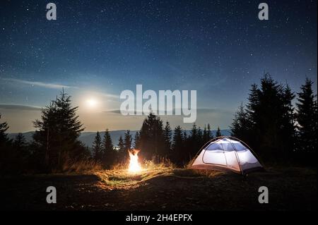 Weißes Zelt auf einem Berghang in der Nähe eines brennenden Lagerfeuers am Wald. Mondschein im magischen Sternenhimmel Übernachtung in den Bergen. Konzept von Reisen, Wandern und Nachtcamping. Stockfoto