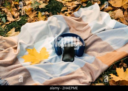 Smartphone-Mockup, Kopfhörer, gelbes Ahornblatt auf Decke im Herbstparkhintergrund. Audio-Technologie-Apps, Meditations-Anwendung, Herbststimmung Musik Stockfoto
