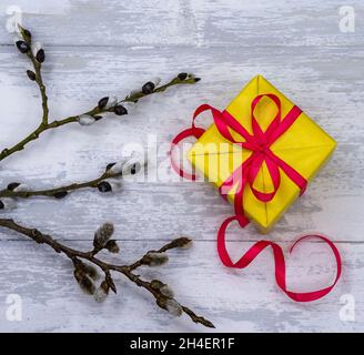 Weidenzweige und ein Geschenk. Es gibt ein gelbes Geschenk auf einem grauen Hintergrund. Geschenkbox mit rotem Band. Stockfoto