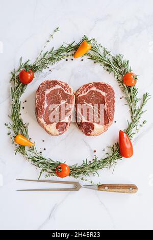 Roh zwei Ribeye-Steaks in der Mitte eines Weihnachtskranzes aus Rosmarin Thymian-Kirschtomaten und Paprika auf einer weißen Marmoroberfläche Stockfoto