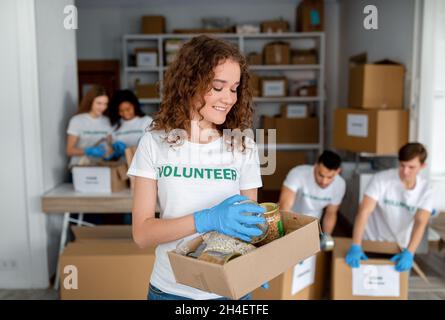 Portrait einer glücklichen Freiwilligen-Frau, die Spendenbox mit Lebensmitteln und Dosen hält, diverse Kollegen, die Produkte verpacken Stockfoto