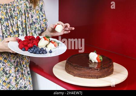 Frau, die in einer roten Küche einen Käsekuchen mit Schokolade und Erdbeeren schmückt Stockfoto