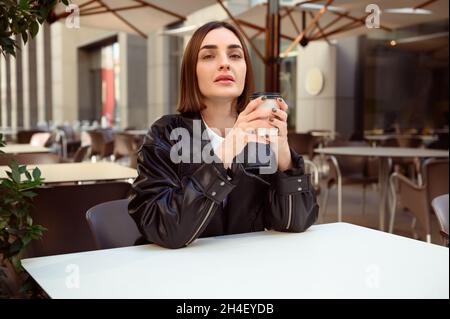 Lifestyle-Porträt einer gut gepflegten Brünette europäische Frau, mit Pappkaffee Blick auf die Kamera, während sie sich auf der Sommerterrasse von außen Kalb Stockfoto