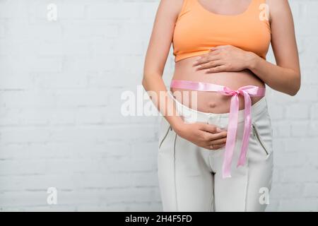 Teilansicht einer Schwangeren mit Satinband auf dem Bauch, das in der Nähe einer weißen Wand steht Stockfoto