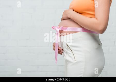 Verkürzte Ansicht einer Schwangeren mit Satinband am Bauch, der in der Nähe einer weißen Wand steht Stockfoto