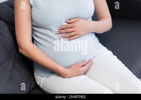 Teilansicht einer Schwangeren, die sich am Bauch umarmt, während sie auf der Couch sitzt Stockfoto