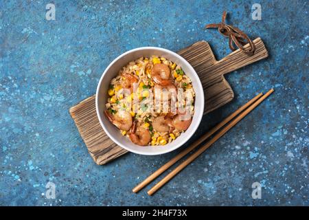 Draufsicht auf eine Schüssel mit chinesischem Reis, leckeren Garnelen, gelbem Mais, Hühnereiern und grünen Zwiebeln auf Holzbrett und blauem Betonhintergrund Stockfoto