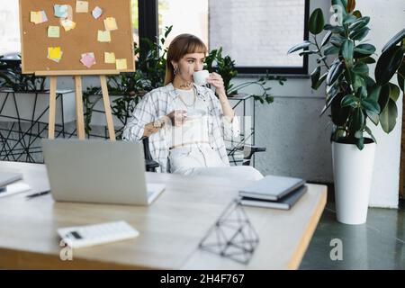 Stilvolle tätowierte Frau, die Kaffee trinkt, während sie am Schreibtisch neben einem verschwommenen Laptop sitzt Stockfoto