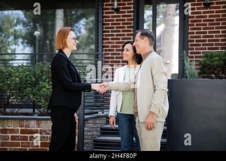 Happy realtor in einer Brille, die sich die Hände schüttelt, während ein Mann in der Nähe der afroamerikanischen Frau und des modernen Hauses steht Stockfoto