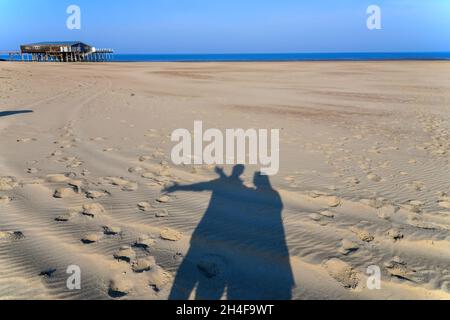Schatten-Selfie von Menschen, die an einem breiten Strand in den Niederlanden gegen einen klaren blauen Himmel bewaffnet sind; Menschen-Schatten reisen um die Welt Stockfoto