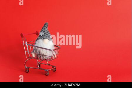 Spielzeug-Gnom im Mini-Einkaufswagen auf rotem Hintergrund mit Kopierfläche Stockfoto