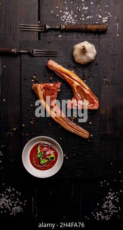 Rohe Lammrippen. Halal-Hammelfleisch. Lammkarree auf dunklem Hintergrund. Salz, Gewürze, Knoblauch, Sauce und Gabeln. Draufsicht. Vertikale Aufnahme. Speicherplatz kopieren Stockfoto