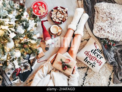 Foto über dem Kopf einer Frau, die ein weihnachtsgeschenk hält, warme Socken trägt und am weihnachtsbaum sitzt Stockfoto