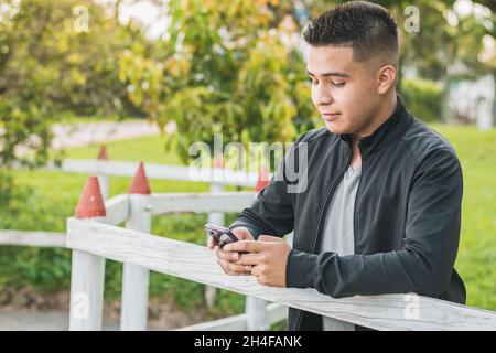 Unternehmungslustiger junger Mann neben dem weißen Holzzaun mit seinem Handy in den Händen, der die Konten seines Unternehmens verwaltet, glücklich über seine Leistungen, wi Stockfoto