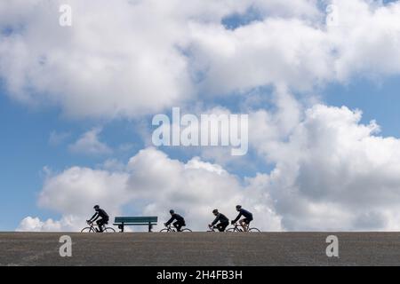 Zeeland, Niederlande-Mai 2021: Blick aus der Nähe auf eine leere Parkbank mit einer Gruppe von Radfahrern, die auf einem der asphaltierten Deiche vorbeifahren Stockfoto