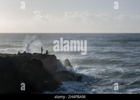 Zwei Sportfischer mit Stangen, die auf einem Felsvorsprung am Atlantischen Ozean stehen, mit rauer See und Wellen, die sich einrollen, wobei sich das Spray an der Küste aufspüllt Stockfoto