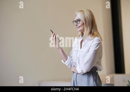 Zu Stoßzeiten. Stilvolle reife blonde Frau in Brille und Business-Kleidung lächeln und sprechen am Telefon, stehen drinnen Stockfoto