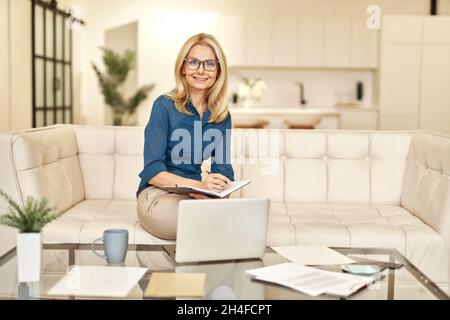 Reife Geschäftsfrau in legerer Kleidung lächelt der Kamera zu und arbeitet von zu Hause aus, während sie auf einer Couch vor dem Laptop in einer modernen Wohnung sitzt Stockfoto