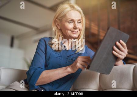 Entspannte, reife Frau, die mit einem digitalen Tablet-pc Nachrichten scrollt und sich zu Hause auf der Couch in einer modernen Wohnung entspannt Stockfoto