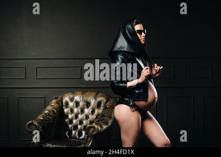 Eine Schwangere in schwarzer Kleidung, Brille und Kopftuch steht mit einem großen Bauch im Studio auf dunklem Hintergrund. Stockfoto