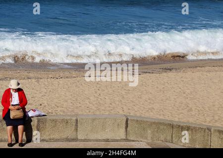 Eine Dame (keine Erkennbarkeit) mit einem roten Blazer, schwarzem Rock und weißem Hut sitzt mit einer Handtasche auf dem Schoß auf einer Strandmauer. Stockfoto