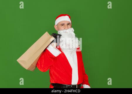 Ein erwachsener kaukasischer Mann, der als Weihnachtsmann gekleidet ist, trägt eine Einkaufstasche aus recyceltem Papier. Der Hintergrund ist grün. Stockfoto