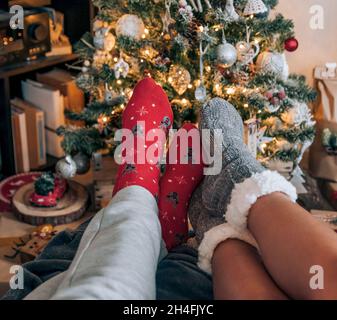 Persönliche Perspektive eines Paares, das kuschelige Socken vor einem geschmückten Weihnachtsbaum trägt. Stockfoto