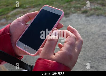Eine Frau hält ein Mobiltelefon in zwei Händen auf der Straße. Kühles, aber sonniges Wetter. Dunkler Bildschirm in einem Smartphone. Stockfoto