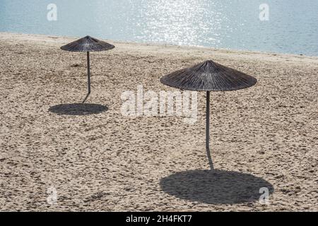 Zwei einsame Schilfschirme an einem leeren Strand am Mittag unter der Sonne. Schatten im Sand. Stockfoto