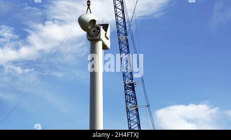 Fernansicht eines Krans, der einen Generator auf einer Windkraftanlage mit einem blauen Himmel im Hintergrund montiert. Stockfoto