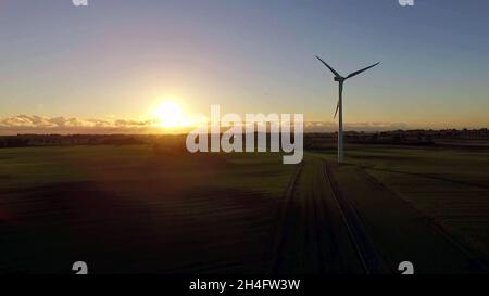 Luftaufnahme von der Drohne und hinter einer Windturbine in der Mitte eines grasbewachsenen Feldes an einem Sonnenuntergangstag. Stockfoto