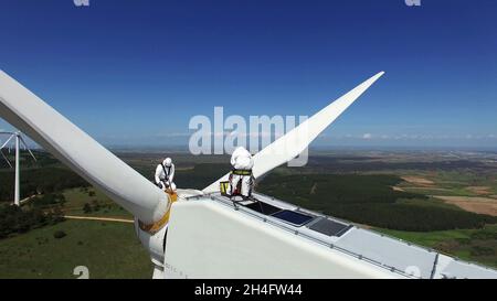 Nahaufnahme einer isolierten Windenergieanlage, bei der Wartungsarbeiter ein Rotorblatt auf dem Generator reparieren. In der Mitte eines grasbewachsenen Feldes mit einem blauen s Stockfoto