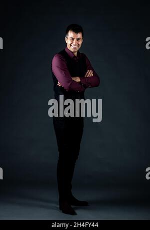 Hübscher Kaukasianer, der mit gekreuzten Armen im Studio auf schwarzem Hintergrund steht und elegante schwarze Hosen und Weste trägt Stockfoto