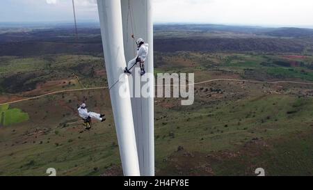 Drohnenansicht von Arbeitern, die einen isolierten Windturbinenmast während Wartungsarbeiten inmitten eines Grasfeldes mit einem bewölkten Himmel im Hintergrund reparieren Stockfoto