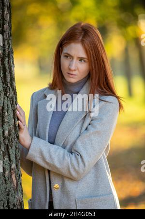 Porträt eines rothaarigen Mädchens im Mantel, das im Herbst hinter einem Baum im Park steht, mit gelben und orangen Blättern auf Baumkronen und Boden Stockfoto