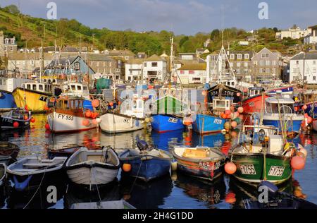 Bunte Fischerboote mit Spiegelungen im Hafen, Geschäften und grünen Hügeln kurz nach Sonnenaufgang in Mevagissey, Cornwall, England Stockfoto